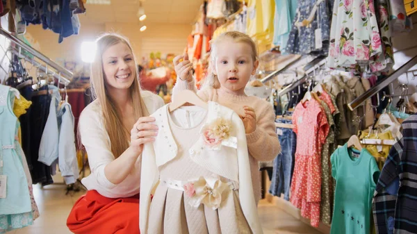 Mujer joven rubia con hija pequeña comprando vestido de niños en la tienda de ropa — Foto de Stock