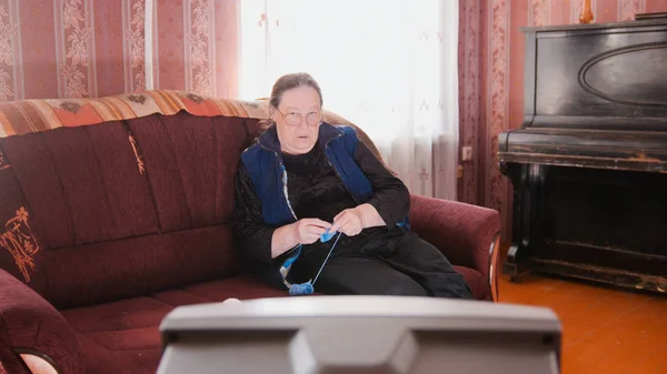 Старушка дома на диване - пожилая женщина смотрит телевизор и вяжет шерстяные носки — стоковое фото