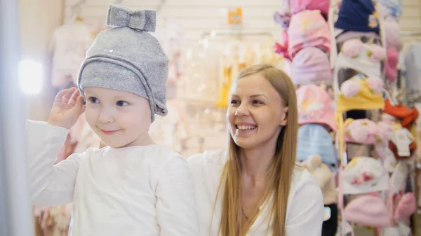 Tienda de vestir para niños - pequeña niña rubia con madre haciendo compras y comprando sombrero — Foto de Stock