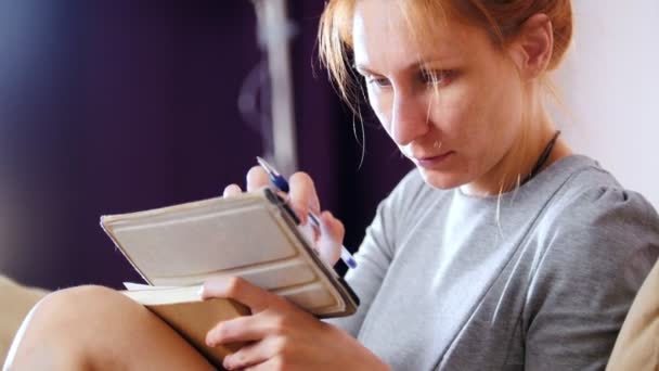 Portrait d'une jolie femme assise à la maison sur un canapé et travaillant avec un gadget et un cahier — Video