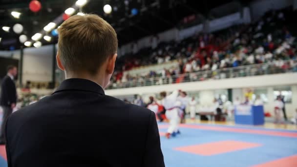 Concursos de arte marcial - hombre entrenador-juez mirando a los adolescentes de karate luchando — Vídeo de stock