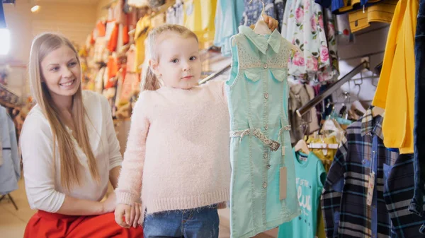 Madre con hija rubia comprando sudadera - ropa para niños en la tienda — Foto de Stock