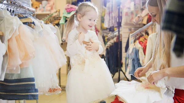 Compras para niños - linda niña con mamá comprando vestido en la tienda de ropa para niños — Foto de Stock