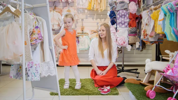 Chicas de compras - niña rubia con mamá elige vestido de moda en la tienda de ropa para niños — Foto de Stock