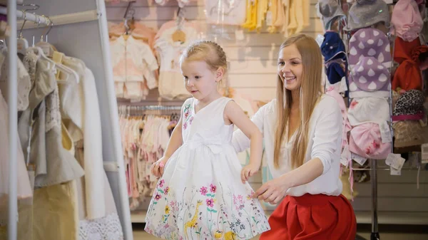 Compras para niños - linda niña con mamá comprando vestido en la tienda de ropa para niños — Foto de Stock