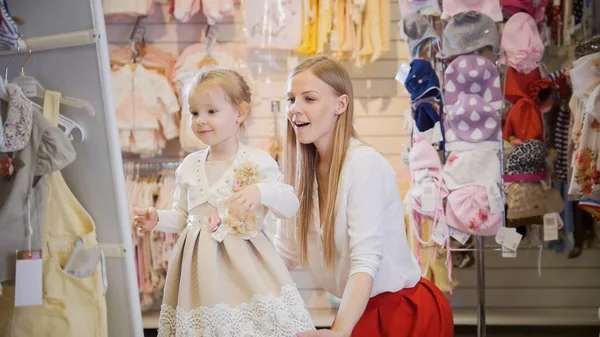 Shopping for kids - blonde little girl with mommy chooses dress in store of kids clothes — Stock Photo, Image