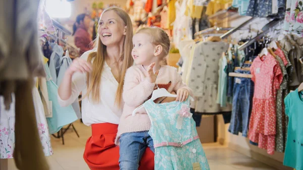 Madre joven con su pequeña hija divirtiéndose en la tienda de ropa para niños — Foto de Stock