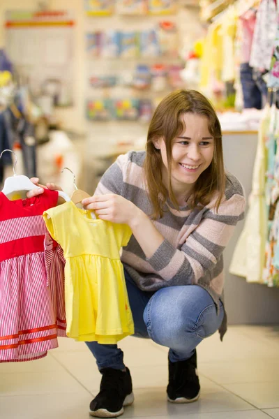 Madre muestra vestidos para su hija - niña en tienda de ropa para niños — Foto de Stock