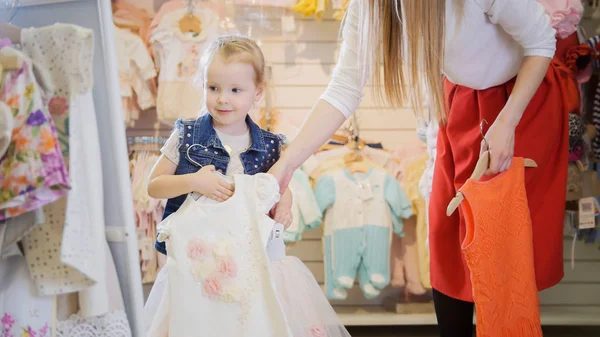 La chica sostiene en sus manos perchas con vestidos delante de un espejo en una tienda de ropa para niños — Foto de Stock