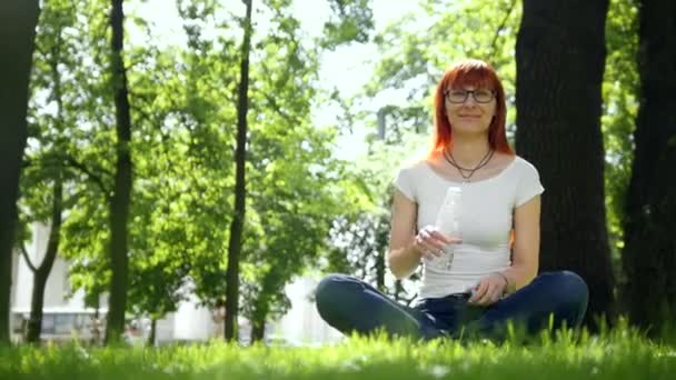 La chica pelirroja bebiendo agua sentada en la hierba en el parque — Vídeos de Stock