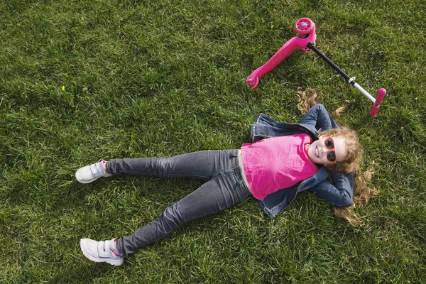 Mosolygó gyermek lány visel farmer kabát feküdt a zöld fű, rózsaszín robogó — Stock Fotó