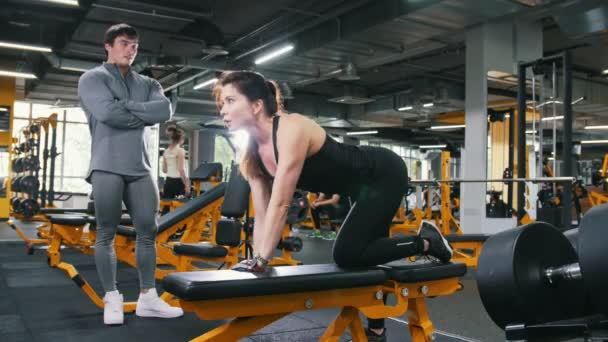 Женщина-спортсмен поднимает гантели в тренажерном зале - тренер наблюдает за тренировками — стоковое видео