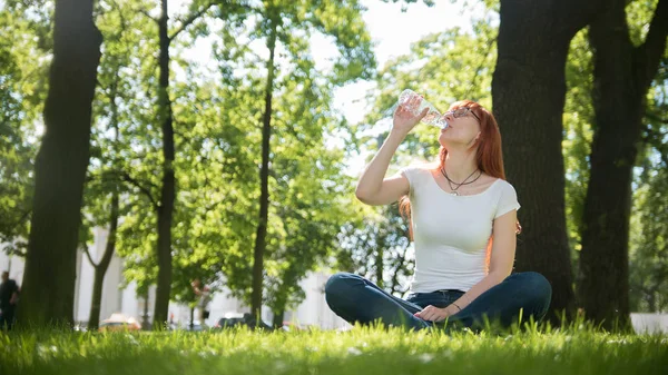 Nő ül a fűben, nyáron szemüveg park és a ital víz — Stock Fotó