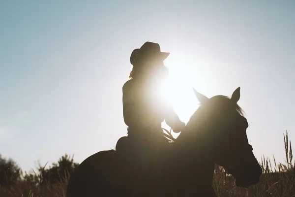 Silhouette d'une femme en chapeau de cow-boy chevauchant un cheval - coucher ou lever de soleil, horizontal — Photo