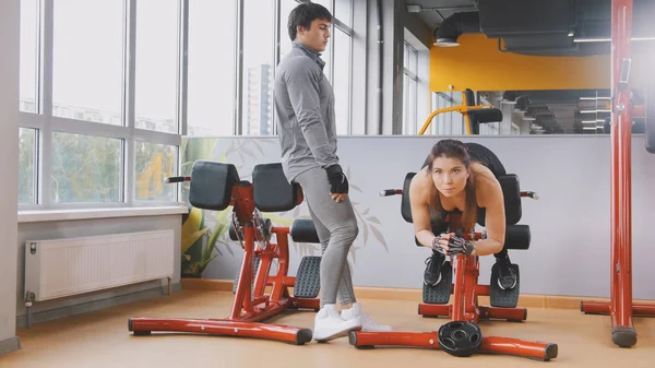 Mulher fitness jovem exercitando abdominal no ginásio - homem muscular olhando para ela — Fotografia de Stock