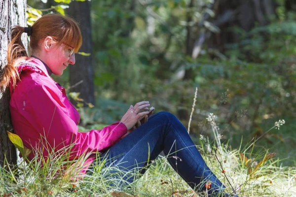 Junge Frau in pinkfarbener Jacke nutzt Smartphone im Herbstwald - waagerecht — Stockfoto