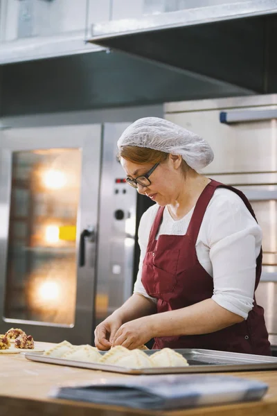 Vuxen kvinna i glasögon och förkläde bakar kakor i bageriet — Stockfoto