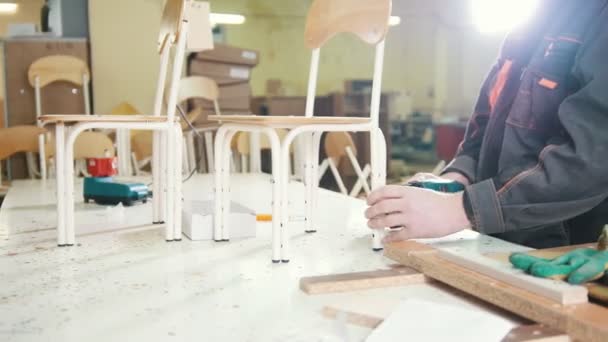 Рабочий плотник прикручивает ножку стула электрической отверткой на мебельной фабрике — стоковое видео