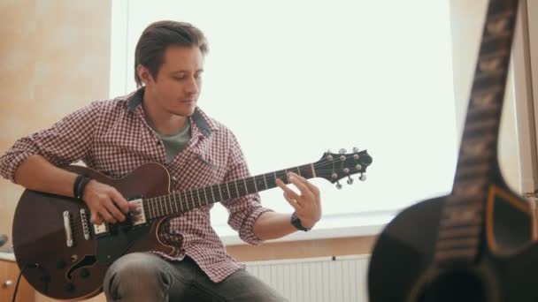 Jovem músico atraente compõe música na guitarra e toca, outro instrumento musical em primeiro plano — Vídeo de Stock