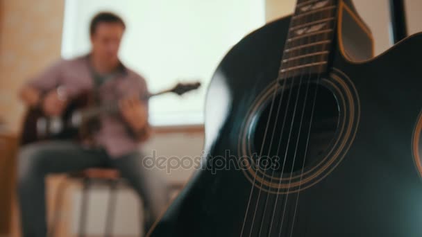 Joven músico atractivo hombre compone música en la guitarra y toca, otro instrumento musical en primer plano, concepto borroso — Vídeo de stock