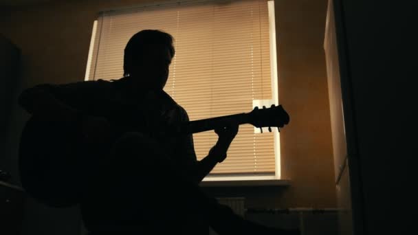 Silhueta escura de jovem músico atraente compõe música na guitarra e toca, outro instrumento musical em primeiro plano, borrado — Vídeo de Stock