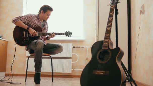 Vonzó fiatal férfi zenész komponál zenét a gitár, és játszik a konyhában, a többi hangszer az előtérben, — Stock videók
