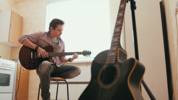 Jonge man componeert muziek op de gitaar en speelt in de keuken, andere muziekinstrument op de voorgrond, — Stockvideo