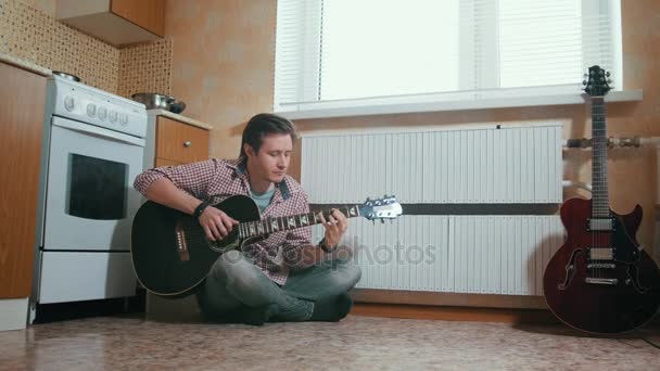 Joven músico toca la guitarra sentado en el suelo en la cocina — Vídeos de Stock