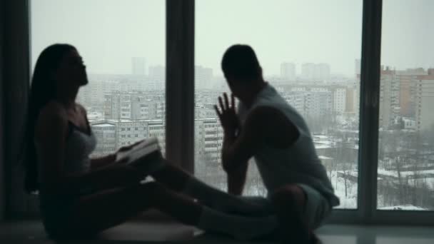 Молодая счастливая пара сидит на подоконнике, девушка читает книгу и улыбающийся парень смотрит в окно — стоковое видео