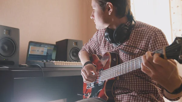 Jovem músico compõe e grava trilha sonora tocando guitarra usando computador, fones de ouvido e teclado — Fotografia de Stock