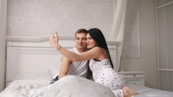 Happy cheerful loving couple making selfie in bed, young attractive guy and girl sitting in pajamas — Stock Video