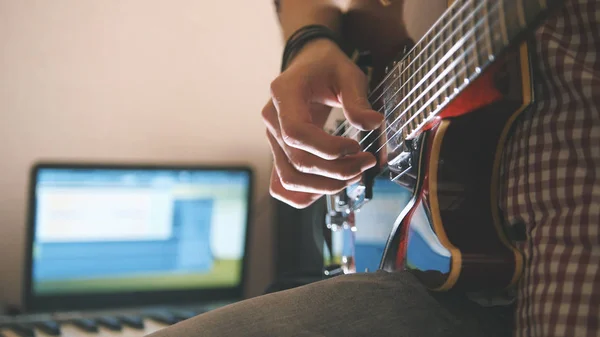 Joven músico masculino compone y graba banda sonora tocando la guitarra usando ordenador, auriculares y teclado — Foto de Stock