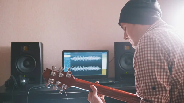 Молодий чоловік у капелюсі музикант пише і записує саундтрек, граючи на гітарі, використовуючи комп'ютер, навушники та клавіатуру — стокове фото