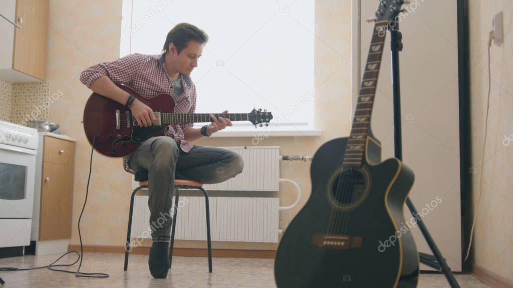 Young man musician composes music on the guitar and plays in the kitchen, other musical instrument in the foreground,