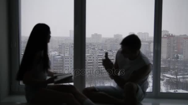 Силует молодої щасливої пари, що сидить на підвіконні, дівчина читає книгу і усміхнений хлопець фотографує вид з вікна — стокове відео