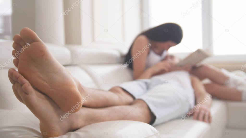 Happy young attractive couple are together on the couch, the girl reads a book, the guy lies on her lap