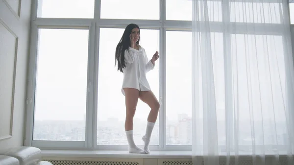 Bella giovane donna in camicia bianca da uomo e calzini che camminano sul davanzale della finestra, ridendo e parlando al telefono felicemente — Foto Stock