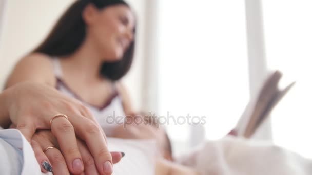 Alliances sur les mains d'un heureux jeune couple attrayant sur le canapé, la fille lit un livre, le gars se trouve sur ses genoux — Video