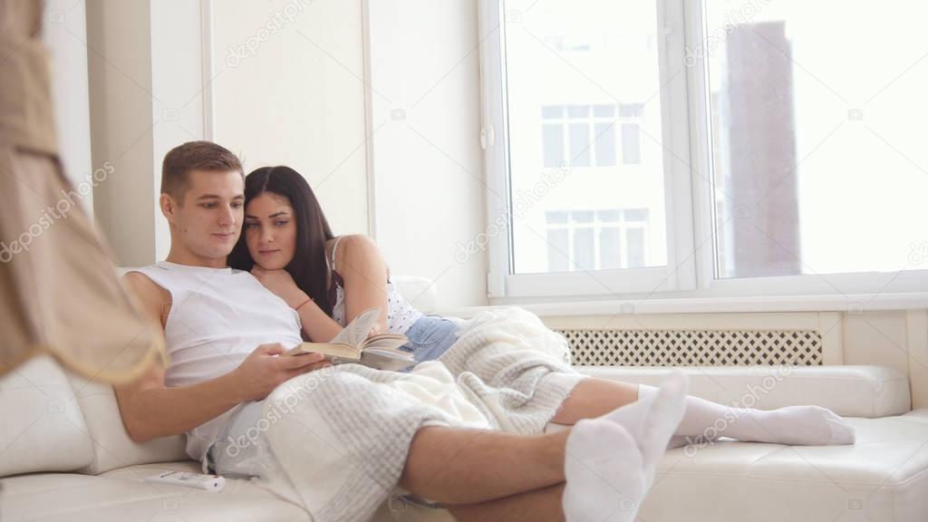 Happy young attractive couple are together on the couch, the girl reads a book, the guy lies on her lap, theyre holding hands