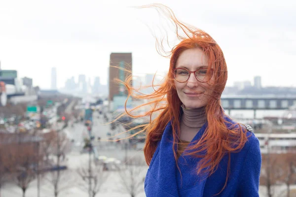 Cute red-haired woman in a blue coat and glasses standing on the background of the big city, her hair fluttering in the wind