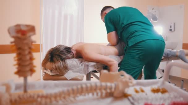 Manueller Therapeut massiert eine junge Frau, die auf einem Massagetisch liegt, und dehnt ihre Gelenke mit seinem Ellbogen, dem Wirbelsäulenmodell und Drogen im Vordergrund — Stockvideo