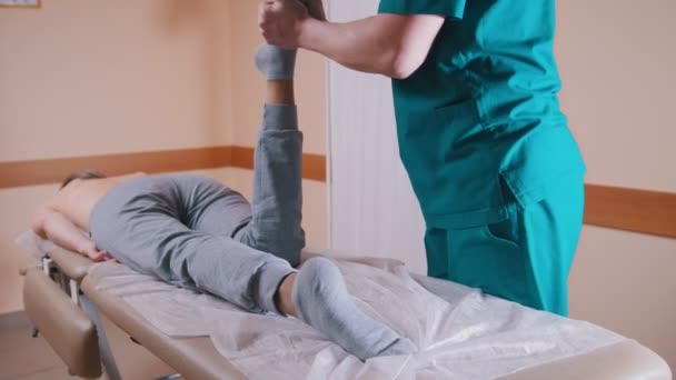 Хиропрактик массирует молодую женщину, лежащую на массажном столе, растягивает и сгибает ноги и колено, крупным планом ног — стоковое видео