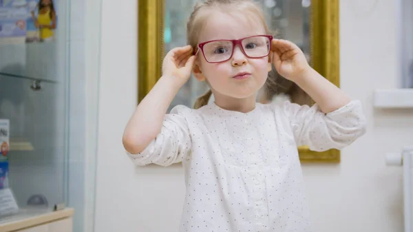 Klein meisje probeert mode medische bril in de buurt van spiegel - winkelen in oogheelkunde kliniek — Stockfoto