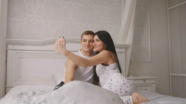 Счастливая любящая пара делает селфи в постели, молодой привлекательный парень и девушка, сидящие в пижаме — стоковое фото