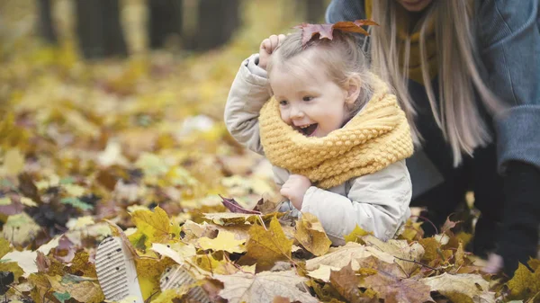 Concepto de familia feliz - hija pequeña con su madre juega con hojas amarillas en el parque de otoño — Foto de Stock