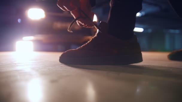 Человек завязывает шнурки на кроссовках на подземной парковке перед фарами автомобиля — стоковое видео