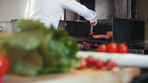 Шеф-повар готовит гриль для приготовления мяса смазывая его маслом на коммерческой кухне, свежие овощи на переднем плане — стоковое видео