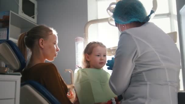 Chica, su madre y el dentista en el consultorio dental, el estomatólogo examinando a la niña con instrumentos especiales — Vídeos de Stock