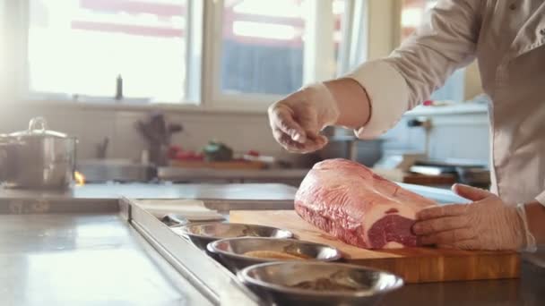 Macellaio aggiungere spezie in un grande pezzo di carne fresca cruda sdraiato su una tavola di legno in una cucina commerciale del ristorante — Video Stock