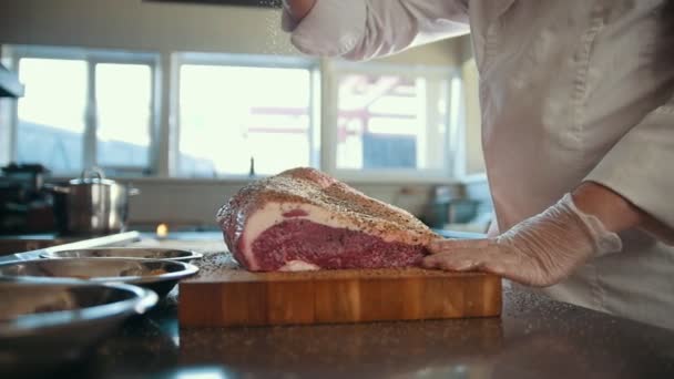Мясник добавляет специи в большой кусок свежего мяса, лежащего на деревянной доске в коммерческой кухне, замедленное движение — стоковое видео